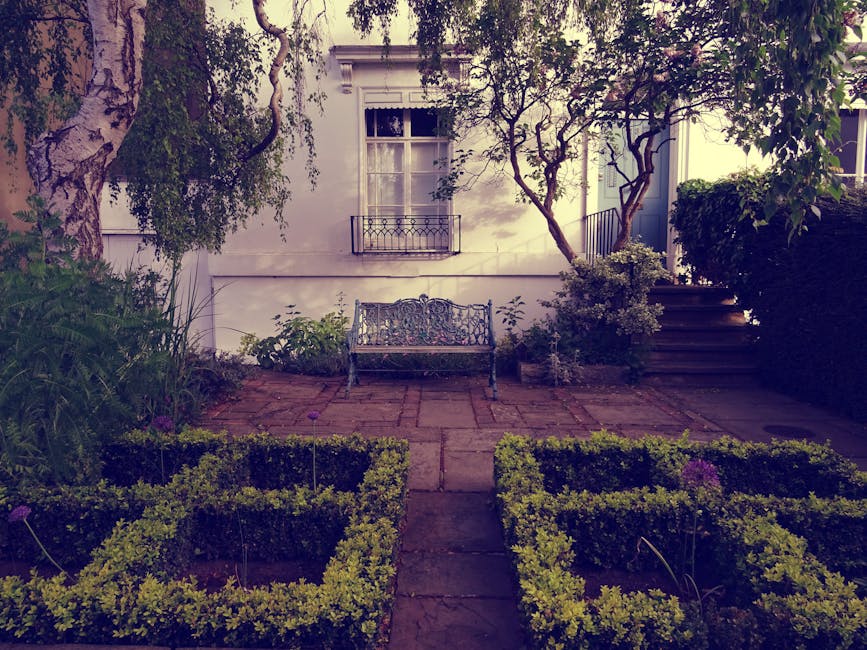 A small landscaped garden area featuring a paved patio with a decorative wrought iron bench positioned against a white exterior wall of a house. The garden is bordered by neatly trimmed boxwood hedges